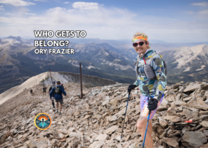 Ory Frazier stands on a rocky mountain overlook wearing a sunglasses and trekking poles, with mountains and a valley behind them.
