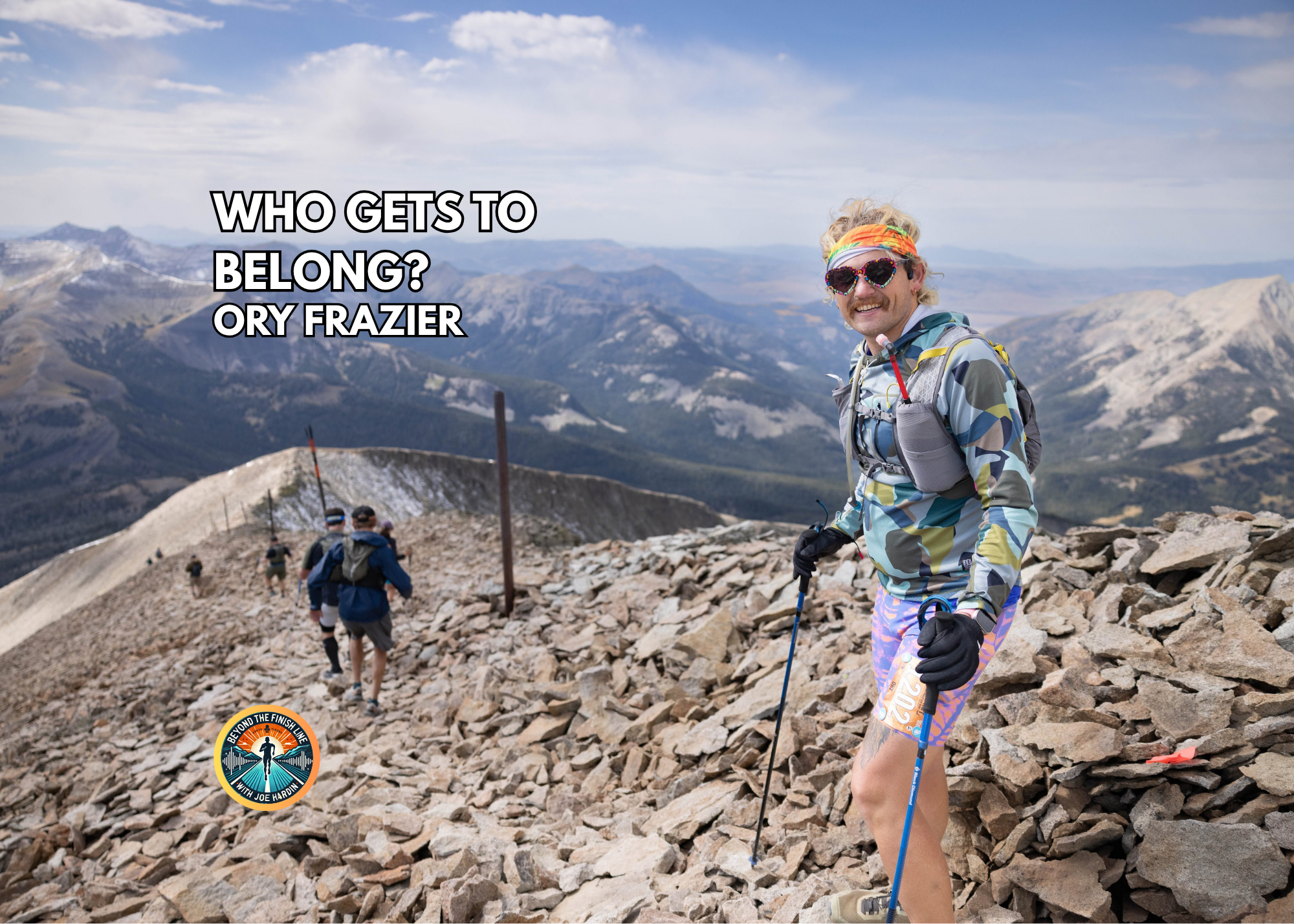 Ory Frazier stands on a rocky mountain overlook wearing a sunglasses and trekking poles, with mountains and a valley behind them.